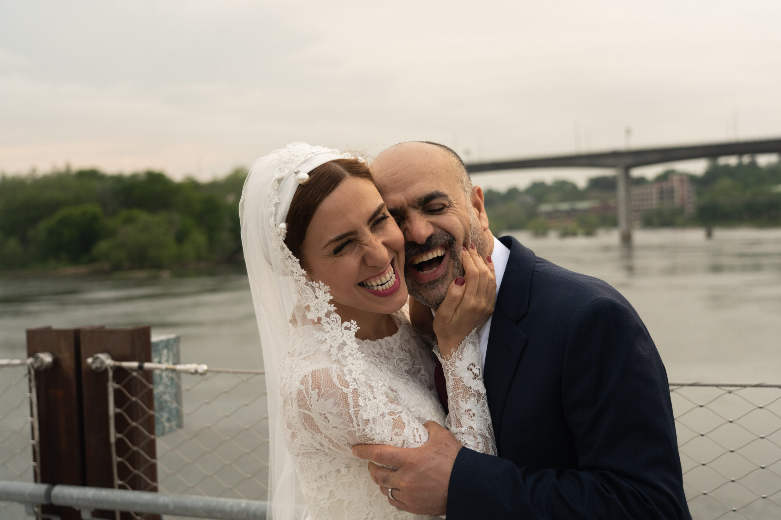 A bride and groom elope on the Potterfield bridge in Richmond, Va. The bride is smiling while squeezing the grooms cheeks with the James River in the background.