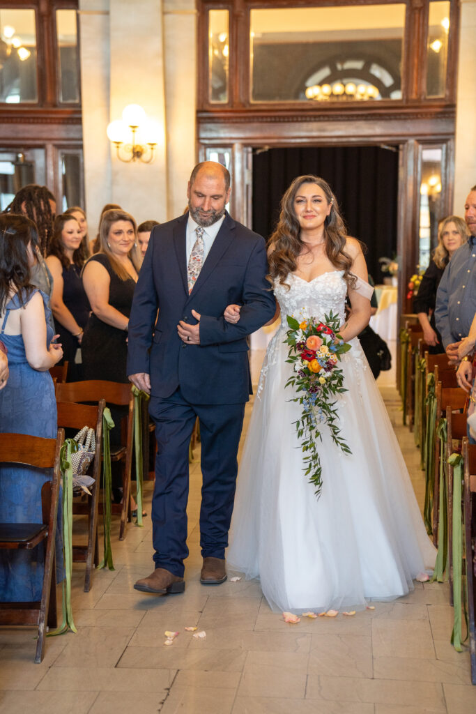 A bride is smiling while holding her wedding boutique. her dad is also smiling while escorting his daughter down the isle as all of the guest smile and watch. 