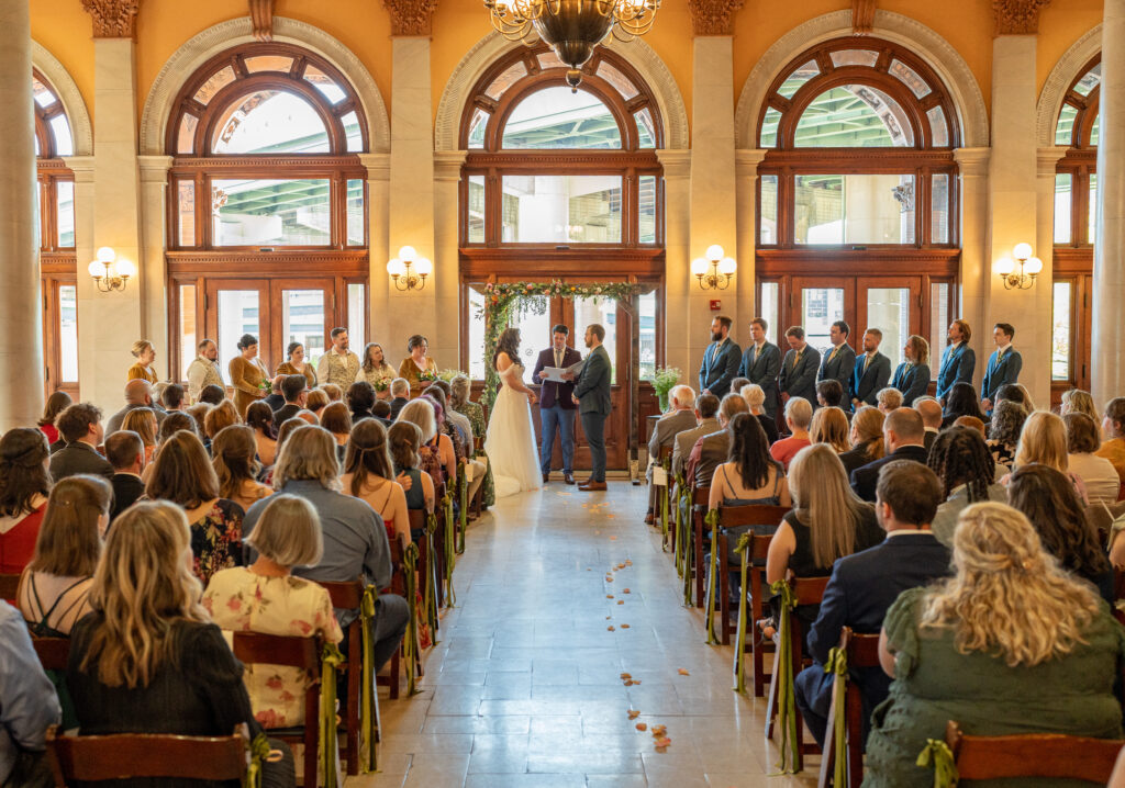 A bride and groom holding hands during their wedding ceremony, guest are sedated and watching  