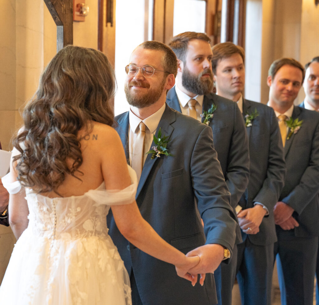 A groom is smiling while holding his brides hand.