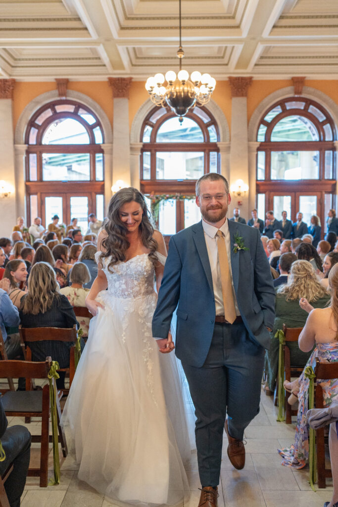 A bride and groom hold hands while walking down the wedding isle at main street station.