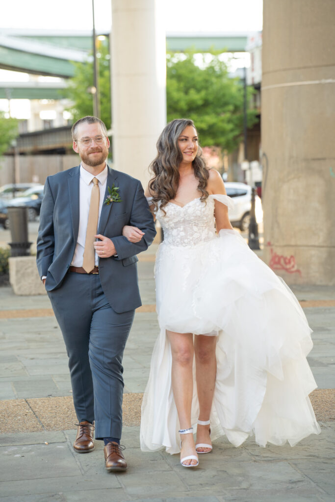 A groom leads his bride across the street while she holds his arm and smiles of pure joy on their wedding day.