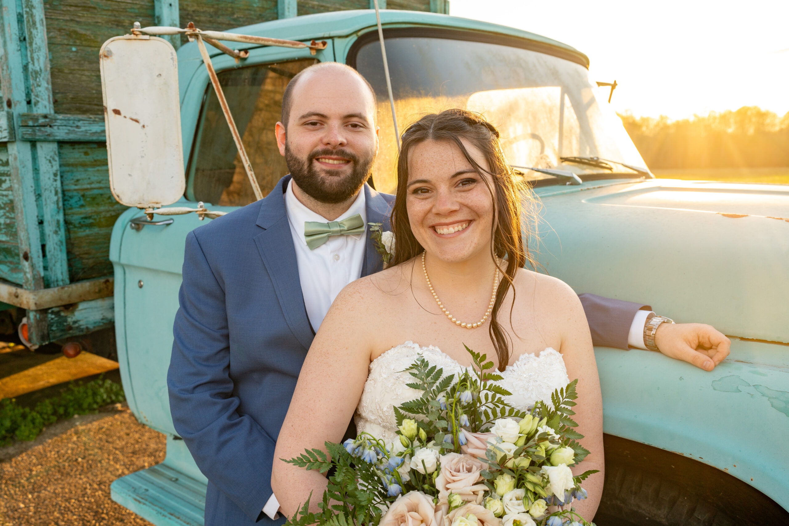 A groom hugs his bride from behind a cousiac manor in Richmond, Virginia. They are standing in front of a vintage farm truck that is blue and the sun is setting behind them during their wedding day photos.