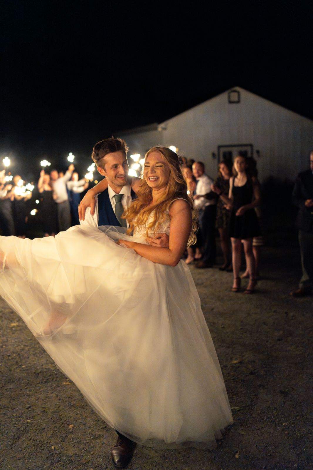 Groom carrying bride during a sparkler exit outside Renback Barn in Rochelle, Virginia, with guests holding sparklers at night